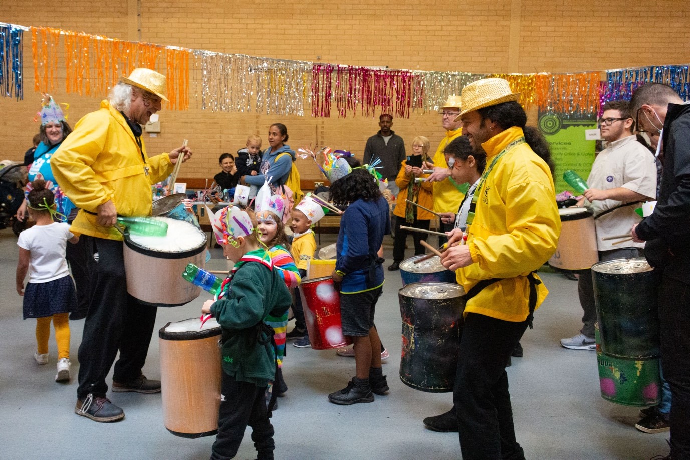 A group of adults and children playing drums in a lively community centre.